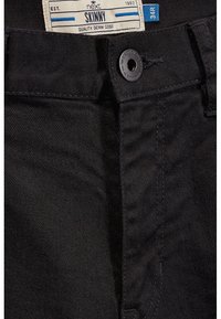 Jeans de mezclilla negros ajustados con cierre de botón estándar y cinturilla. La etiqueta dice "next SKINNY". Textura suave con detalles de costura típicos.
