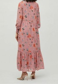 Femme portant une longue robe rose à fleurs avec des manches longues, des poignets froncés et un ourlet à volants, accompagnée de chaussures à talons hauts dorées.