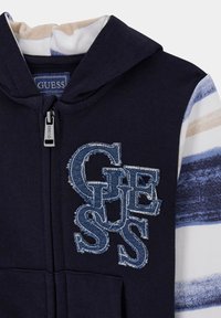 Sweat à capuche bleu marine avec un intérieur rayé contrastant et un grand logo "GUESS" en denim usé sur la poitrine ; comprend une fermeture éclair et une capuche.