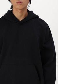 Schwarze Kapuzenjacke aus weichem Stoff, mit Fronttasche, Kordelzugkapuze und lockerer Passform, mit minimalen Nähedetails.