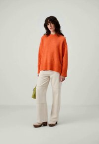 Orange knit sweater with a relaxed fit and ribbed cuffs, paired with cream-colored trousers and brown platform shoes. Green bag in hand.