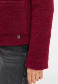 Maglione borgogna con un design in maglia testurizzata e una grande tasca a toppa. Presenta un piccolo tag in metallo vicino all'orlo. Tessuto morbido e vestibilità comoda.
