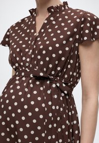 Καφέ φορέμα με λευκές πουά βούλες, γιακά με βολάν, κοντά μανίκια με φτερωτό σχέδιο, κουμπωτό μπροστινό μέρος και ζώνη στη μέση δεμένη σε φιόγκο σε έναν κορμό ανθρώπου.