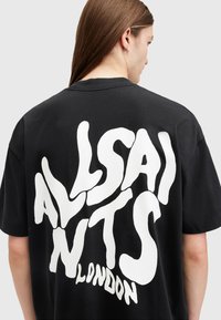 Camiseta de algodón negra con gran texto ondulado en blanco "ALLSAINTS LONDON" en la parte posterior. Mangas cortas y ajuste relajado.