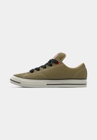 Olive canvas sneakers with black laces, a low-profile design, white rubber sole, and contrasting black and white accents. Red label detail on tongue.