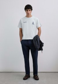 Tânăr cu păr creț și închis la culoare, purtând un tricou grafic gri deschis, pantaloni bleumarin, pantofi maro, ținând o jachetă închisă într-o mână.