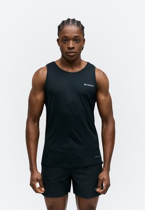 Μυώδες άτομο με πλεξούδες, που φοράει μαύρο αμάνικο τοπ Columbia και μαύρα σορτς, στέκεται μπροστά σε ένα απλό φόντο.