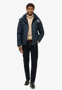 Chaqueta acolchada azul marina con cuello alto y cierre de cremallera, suéter de cuello alto crema, jeans oscuros y zapatos negros. Presenta acentos de logotipo en la chaqueta.