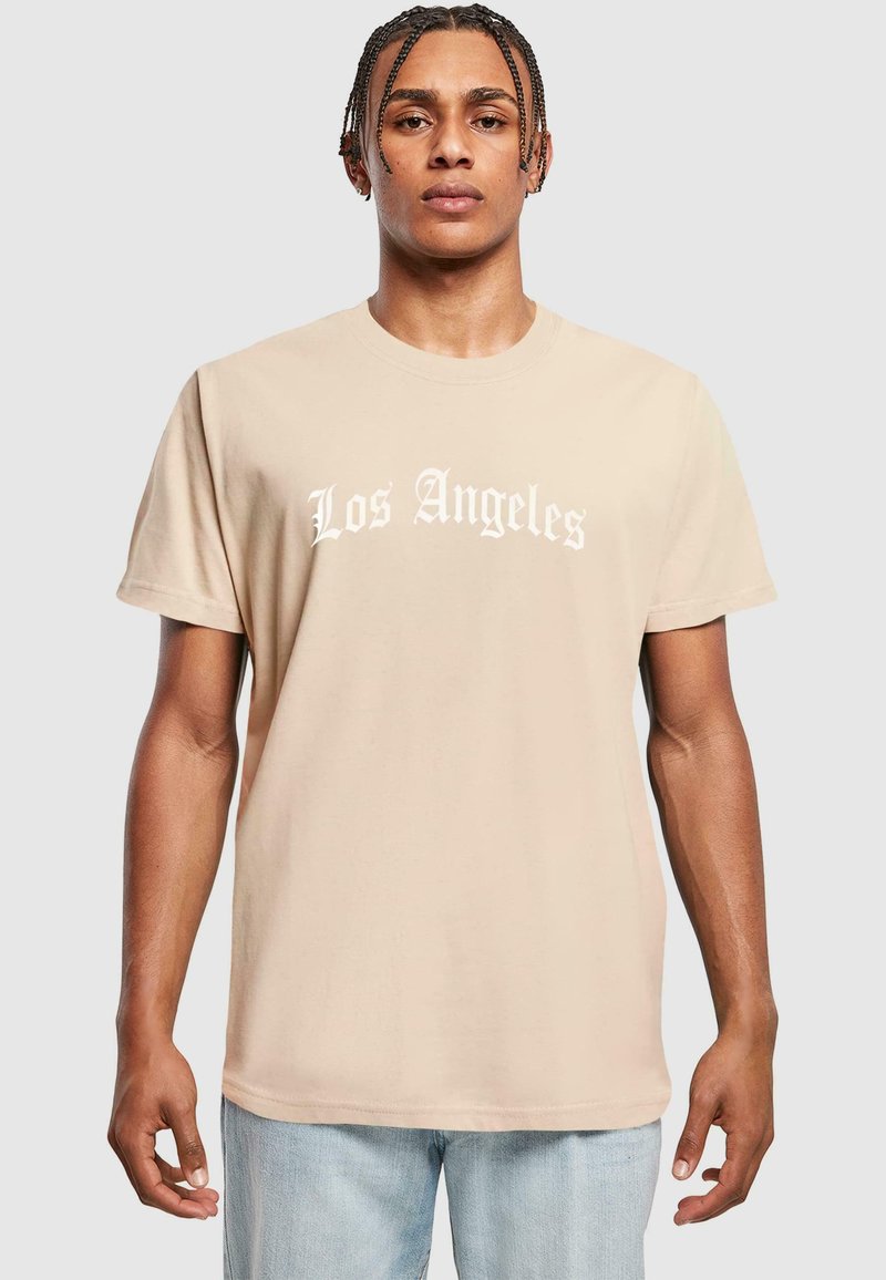 Beige pamut póló, amelyen fehér betűkkel szerepel a "Los Angeles" felirat, rövid ujjú és laza szabású, világoskék farmernadrággal párosítva.