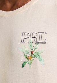 Camiseta de algodón beige con un gráfico floral en verde y rojo con el texto "PIRL" en negro. Cuello redondo con una textura suave.