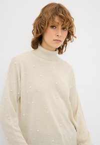 Beige, hoge hals trui met een losse pasvorm. Getextureerd met kleine, ronde, witte versieringen die gelijkmatig over het oppervlak zijn verdeeld.