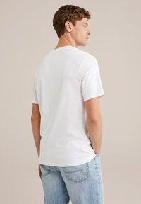 Mlad moški s kodrastimi lasmi, oblečen v enostavno belo majico s kratkimi rokavi in svetlo modre jeans hlače, obrača se na stran proti nevtralnemu ozadju.