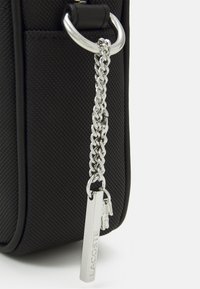 Svart texturerad väska med en silverkedja som accent, en rektangulär tagg präglad med "LACOSTE" och dekorativa charms.