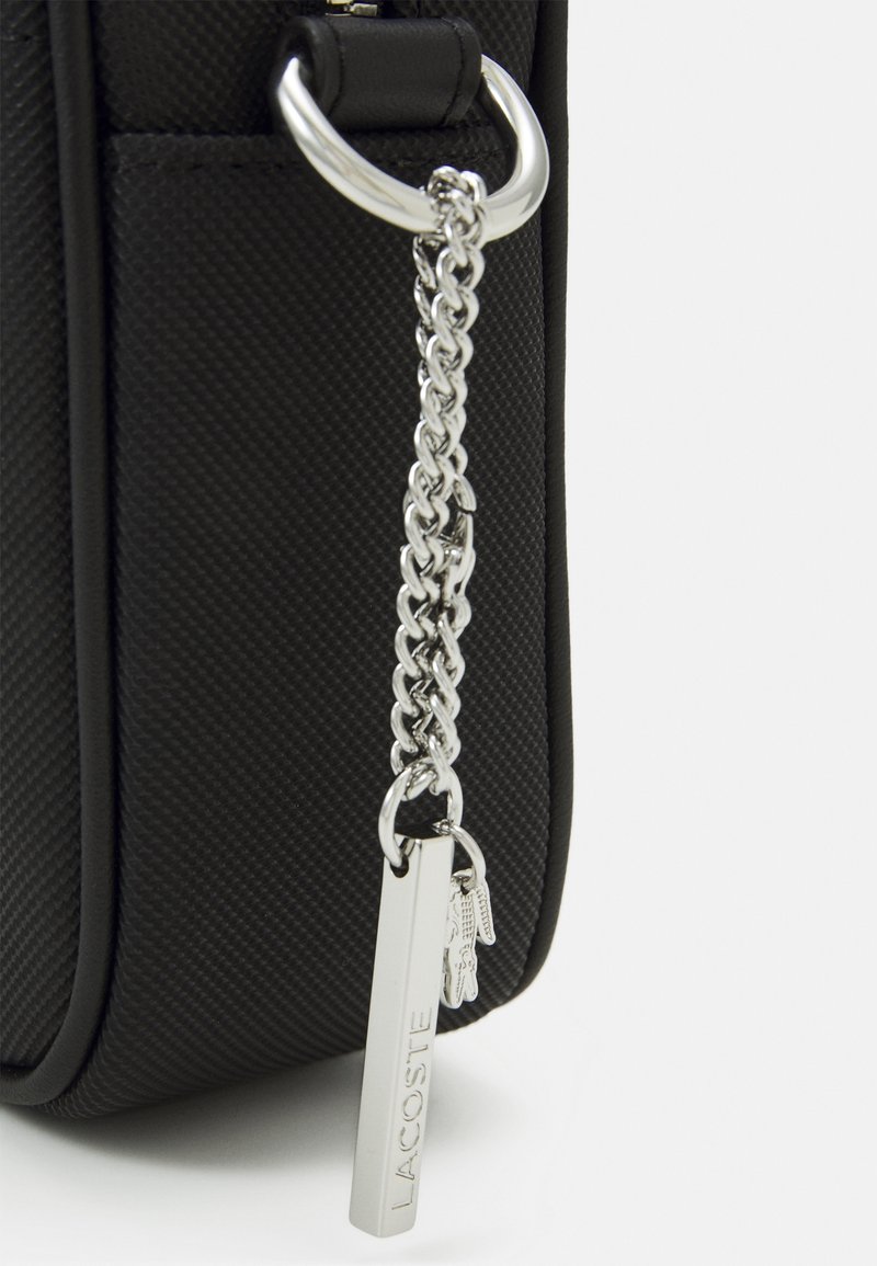 Svart texturerad väska med en silverkedja som accent, en rektangulär tagg präglad med "LACOSTE" och dekorativa charms.