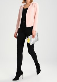 Veste rose pâle à finition texturée, haut noir, jean skinny noir, et bottines noires à bout pointu. Pochette argentée métallique avec un accent feuille.