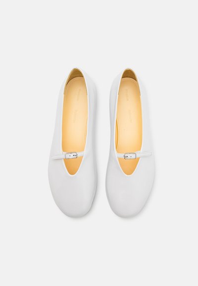 White ballet flats with a rounded toe and a decorative buckle on a strap. Smooth leather exterior and a textured yellow insole.