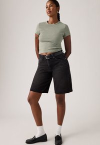 Νεαρή γυναίκα με πλεγμένα μαλλιά, φοράει πράσινη ριγέ κοντομάνικη μπλούζα, μαύρο σορτς μέχρι το γόνατο με ζώνη, λευκές κάλτσες και μαύρα μοκασίνια.