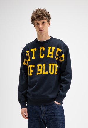 Jeune homme aux cheveux bouclés portant un sweat-shirt bleu marine avec un grand texte jaune et un jean bleu, debout sur un fond blanc.