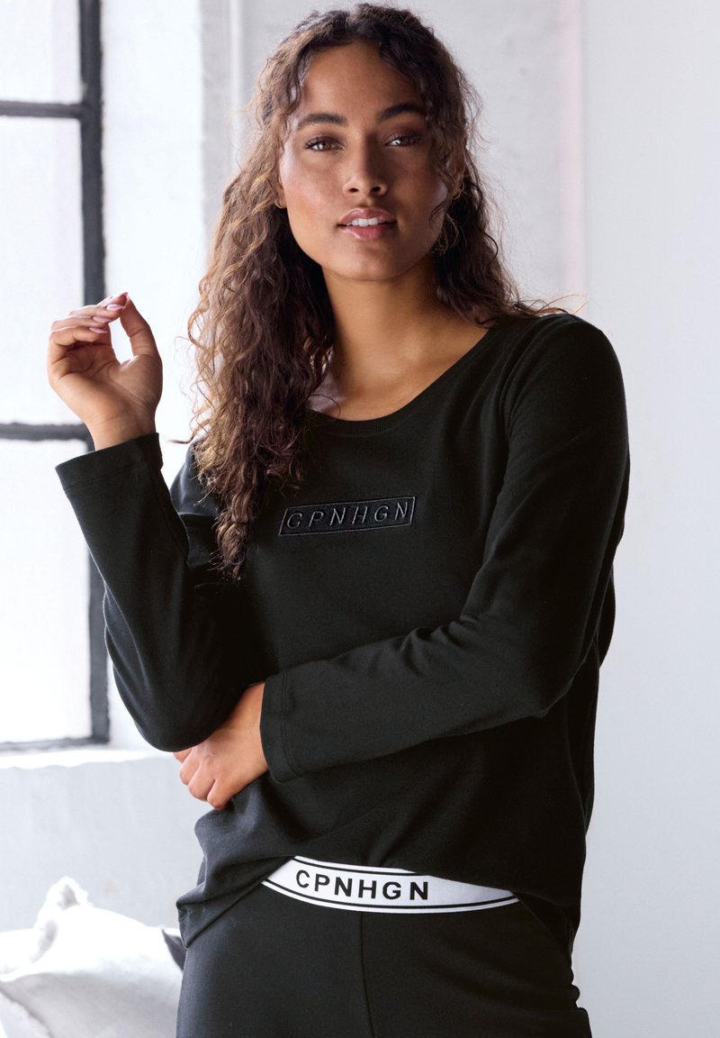 Schwarzes Langarmshirt mit rundem Ausschnitt, versehen mit einem geprägten Markenlogo. Das Model trägt passende schwarze Hosen mit einem weißen Logo-Bund.