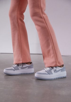 Stopy w szaro-białych sneakersach na platformie w połączeniu z luźnymi różowymi spodniami stojące na szarej betonowej posadzce.