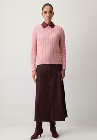 Žena v ružovom svetri s vrkočovým vzorom a bordovým golierom, dlhej bordovej sukni a zodpovedajúcich bordových čižmách na opätku, stojaca pri bielej stene.