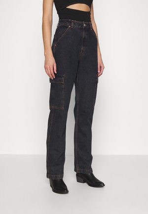 Femme portant un jean cargo noir taille haute avec poches à rabat et des bottines noires à talons, sur un fond blanc uni.