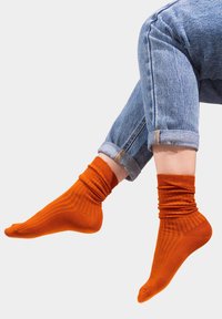 Pomarańczowe bawełniane skarpetki z prążkowaną fakturą i zebranym mankietem, noszone z podwiniętymi niebieskimi dżinsami.