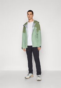 Veste d'extérieur vert clair avec des accents gris, fermeture éclair, et capuche ajustable, associée à un pantalon noir et des chaussures de sport grises.