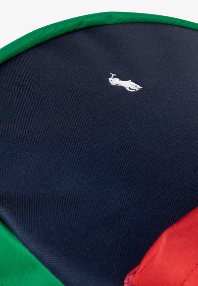 Détail d'un sac à dos en tissu bleu marine avec une bordure verte et une accent rouge. Il présente un petit logo brodé blanc d'un cavalier. Surface texturée.