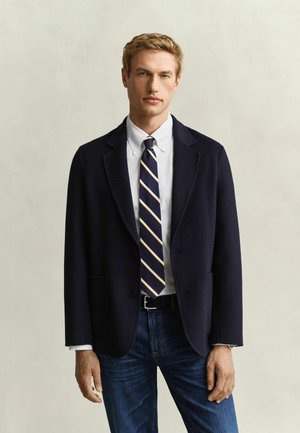 Man die een marineblauwe blazer, wit overhemd, een marineblauw-geel gestreepte stropdas en blauwe jeans draagt, staand tegen een effen lichte achtergrond.