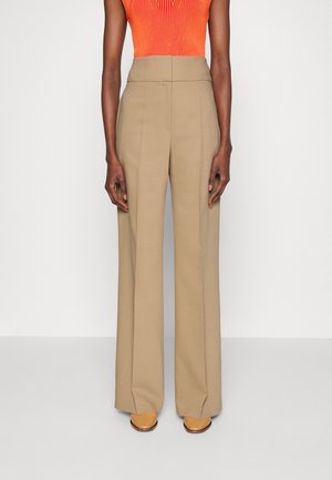 Person iført beige bukser med højt talje og vide ben, orange ærmeløs top og brune spidse sko, stående mod en ensfarvet hvid baggrund.