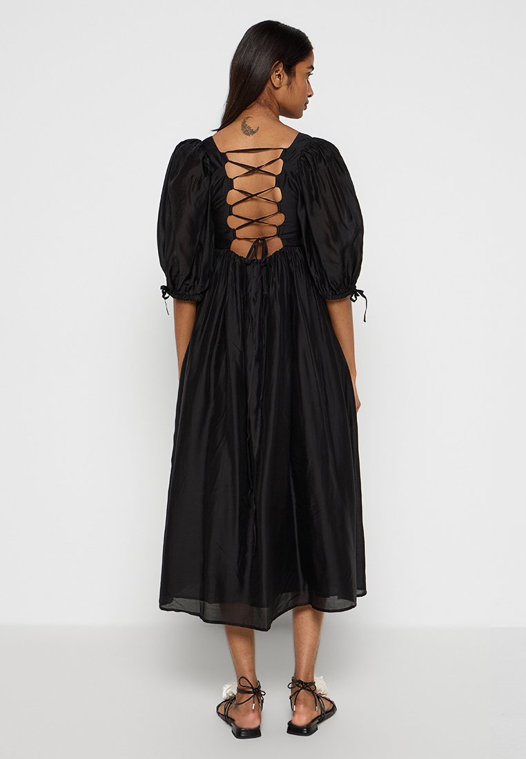 Robe noire mi-longue avec des manches bouffantes, détail de dos ouvert avec lacets, et texture satin fluide. Se noue à l'arrière et est portée avec des sandales plates.