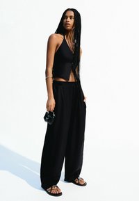 Top court noir, pantalon large noir ample, et sandales à lanières. Accessoires comprenant un petit sac noir. Cheveux longs tressés.