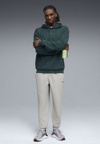 Felpa verde scuro con tasca frontale e cordini; pantaloni della tuta grigi chiari con logo; scarpe da ginnastica grigie e amaranto; tiene in mano una bottiglia verde.