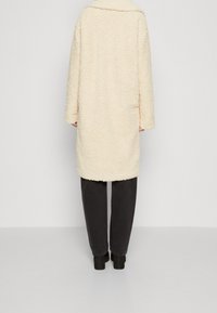 Abrigo de gran tamaño en tono crema, suave y de textura, sin botones y con cuello con muesca, combinado con pantalones oscuros y botas gruesas negras.