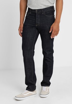 Ein Mann trägt dunkelblaue Slim-Fit-Jeans, ein hellgraues T-Shirt und beige-weiße Sneakers und steht vor einem einfarbigen Hintergrund.