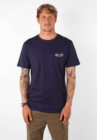 T-shirt en coton bleu marine avec un col rond, des manches courtes et un petit logo floral "OXBOW" sur la poitrine gauche.