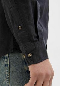 Cămașă din in gri închis, cu manșete cu nasturi și nasturi aurii. Cusături vizibile și un material ușor texturat. Fundal din denim.