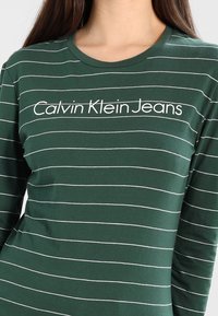 Grünes Langarmshirt mit weißen horizontalen Streifen und rundem Halsausschnitt. Logo "Calvin Klein Jeans" prominent in der Mitte angezeigt.