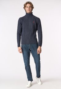 Giovane uomo che indossa un maglione a collo alto di colore blu navy con lavorazione a trecce, jeans blu e sneaker bianche, in piedi contro uno sfondo semplice.