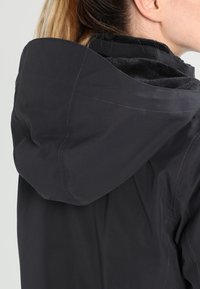 Femme portant une veste à capuche noire avec doublure en polaire, vue de dos et légèrement tournée sur le côté.
