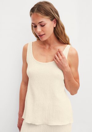 Vrouw met lichtbruin haar draagt een mouwloze, gestructureerde beige top, raakt zachtjes haar schouder aan en kijkt naar beneden tegen een effen lichte achtergrond.