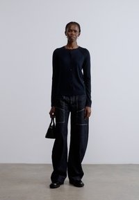 Čierny kardigán na gombíky, skombinovaný so širokými tmavými džínsovými nohavicami s prednými vreckami. Modelka drží malú čiernu kabelku a stojí na neutrálnej podlahe.
