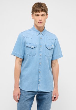 Junger Mann trägt ein hellblaues kurzärmeliges Jeanshemd mit Druckknöpfen und zwei Brusttaschen, kombiniert mit blauen Jeans, vor einem einfarbigen Hintergrund.