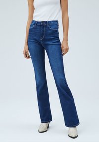 Flared Jeans aus dunkelblauem Denim, mit mittlerer Taille, fünf Taschen, weißen Nähten und Metallknopfdetails. Glatte Textur mit leichtem Stretch.
