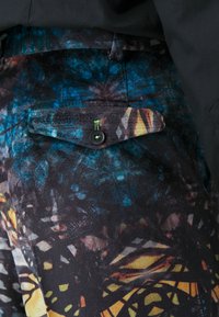 Multi-colored fabric pants featuring an abstract pattern of blue, orange, and black, with a back pocket secured by a black button.