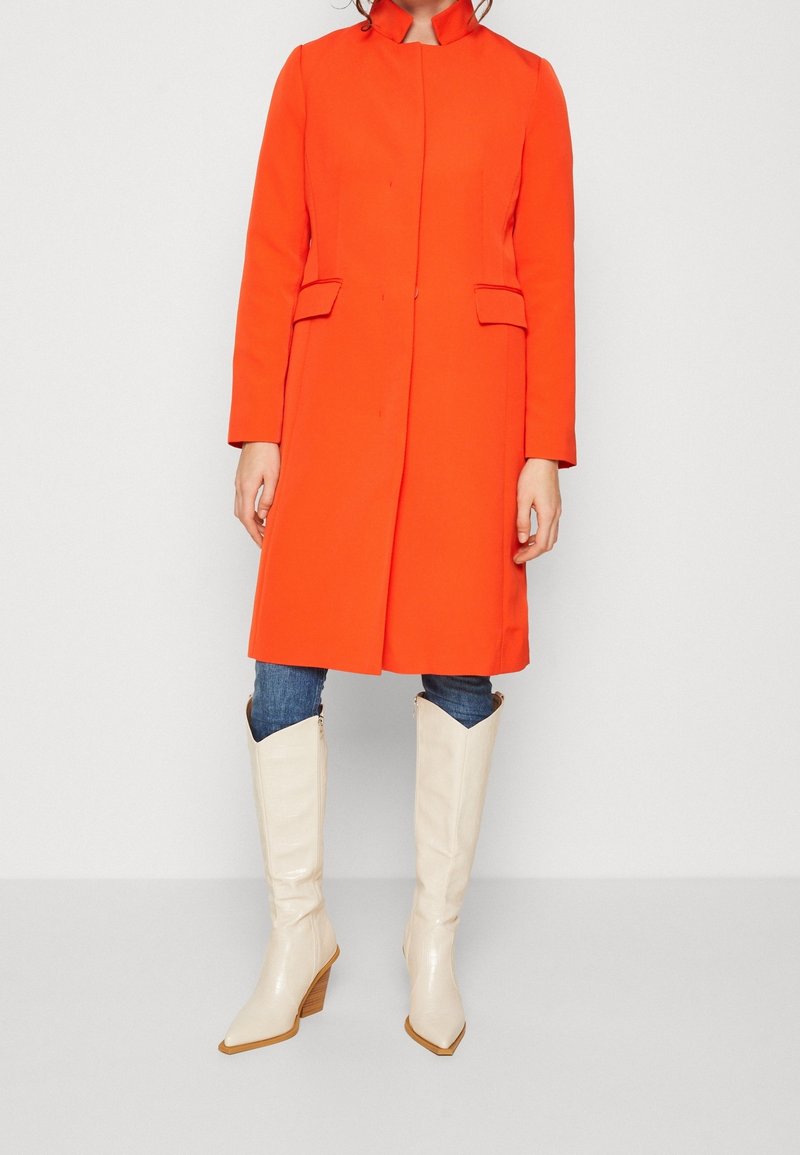 Lys orange, skræddersyet frakke med høj krave, med to forlommer og lige snit, kombineret med hvide, knæhøje støvler med hæl.