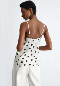 Camisola bianca con pois neri; tessuto leggero, spalline sottili regolabili e chiusura a bottone sul retro. Texture liscia.