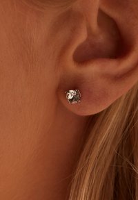 Runde silberfarbene Steckerohrringe mit klarem Kristall, der mit vier Krappen gefasst ist, und nah am Ohrläppchen positioniert. Glatte metallische Oberfläche.