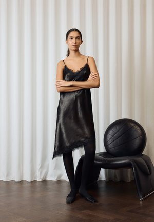 Kvinde står med armene over kors iført en sort satin natkjole, sorte strømpebukser og sorte sko ved siden af en rund sort læderstol og hvide gardiner.